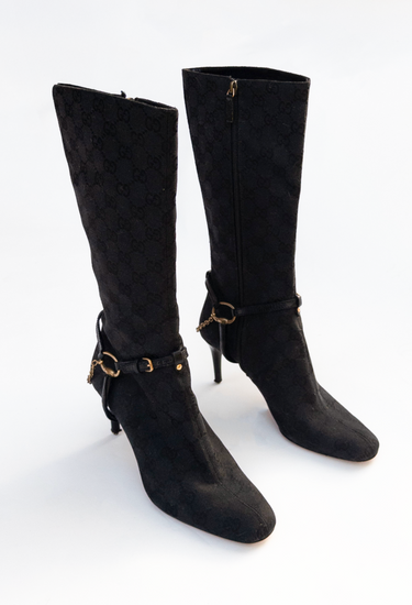 Gucci Tom Ford knee boots in black GG monogram canvas with  with leather harness straps, anchored by gold-tone horse bit hardware and fine chain - angled view